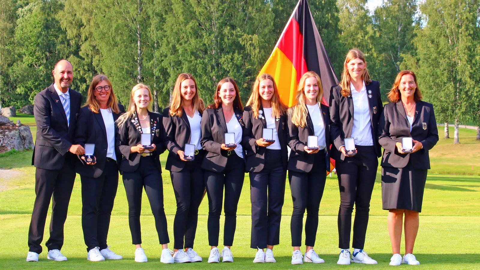 Nationalteam in dunklen Blazern mit Medaillen und Deutschlandfahne in einer Reihe auf dem Grün