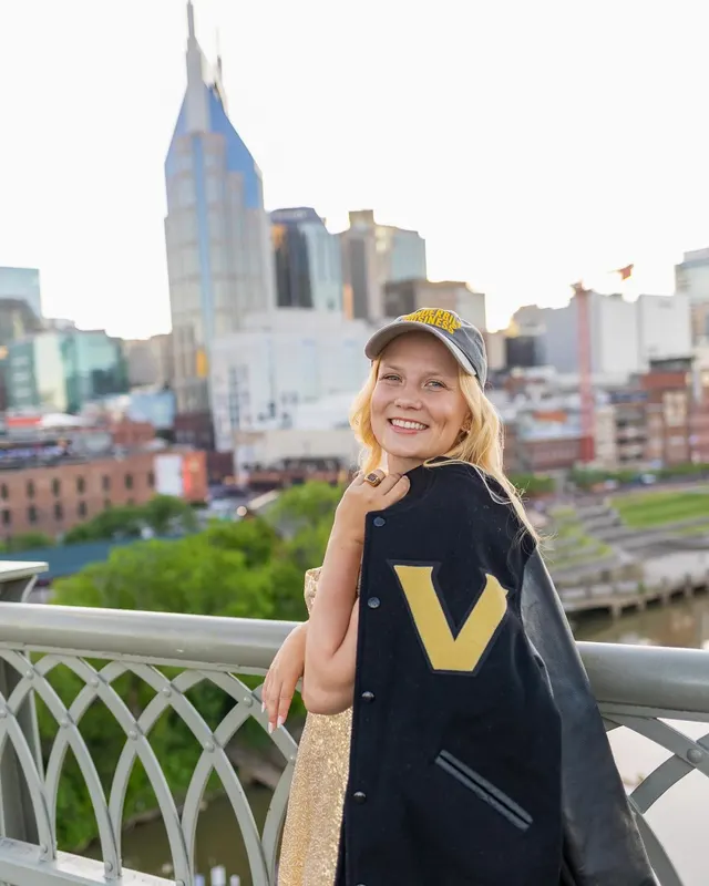 Celina Sattelkau: lacht über die Schulter in Vanderbilt-Jacke und Cap, Stadtkulisse im Hintergrund