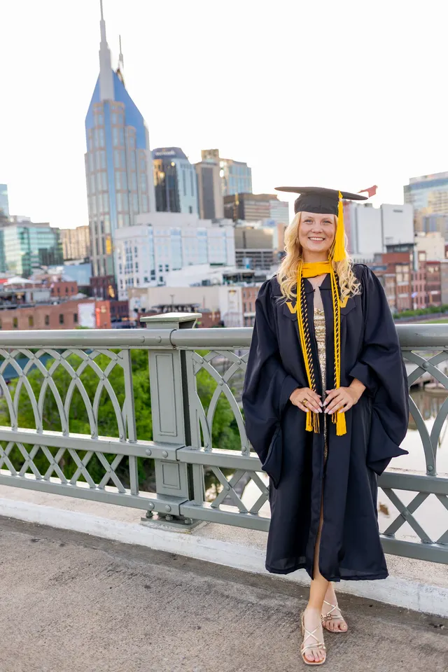 Celina Sattelkau: Ganzkörper im Talar mit Doktorhut und goldener Kordel auf der Brücke, Stadt im Rücken