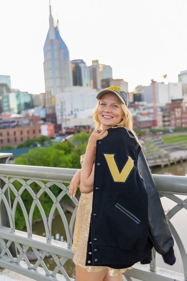 Celina Sattelkau: lacht in Vanderbilt-Jacke und goldener Cap an der Brückenreling, Stadt dahinter